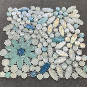 May include: A mosaic tile sheet featuring a floral design. The mosaic is composed of various shapes and sizes of glass tiles in shades of blue, white, and iridescent colors. The design includes a large flower in the center and scattered accents.