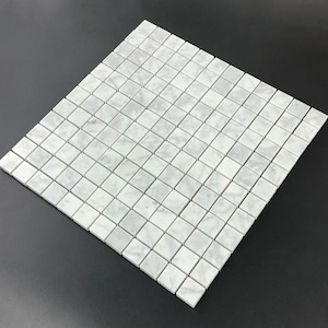 May include: A square sheet of white marble mosaic tiles. Each tile is a small square, creating a grid pattern. The marble has subtle gray veining, adding texture. The tiles are arranged on a dark surface.