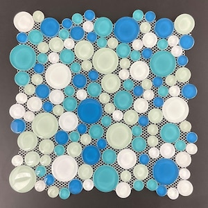 May include: A square mosaic tile sheet featuring a pattern of various sized, round glass tiles. The tiles are in shades of white, light green, turquoise, and blue, set on a white mesh backing. The background is a solid dark gray.