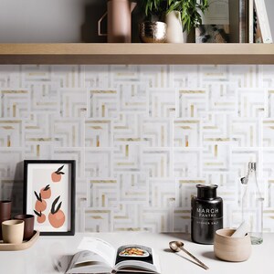 May include: A white kitchen backsplash with a geometric pattern of gold and white tiles. The backsplash is behind a counter with a cookbook, a salt shaker, a mortar and pestle, and a framed print of oranges.