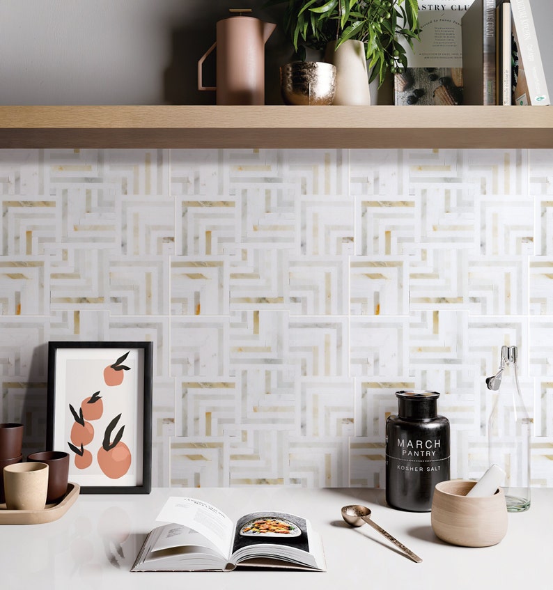 May include: A white kitchen backsplash with a geometric pattern of gold and white tiles. The backsplash is behind a counter with a cookbook, a salt shaker, a mortar and pestle, and a framed print of oranges.