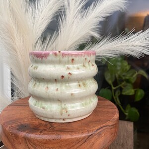 May include: A white ceramic planter with a green glaze and pink rim. The planter has a wavy, textured design and is sitting on a wooden stand.