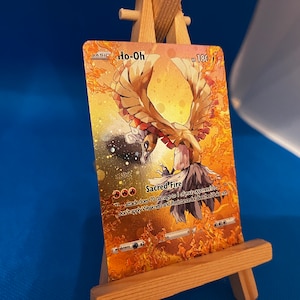 Pu&ograve; includere: Una carta da gioco di Pokemon dorata con Ho-Oh, un Pokemon uccello leggendario con un design arancione e giallo fiammeggiante. Il testo della carta recita "Sacred Fire: Questo attacco infligge 70 danni a 1 dei Pokemon del tuo avversario in gioco. Non applicare Debolezza e Resistenza ai Pokemon in panchina."