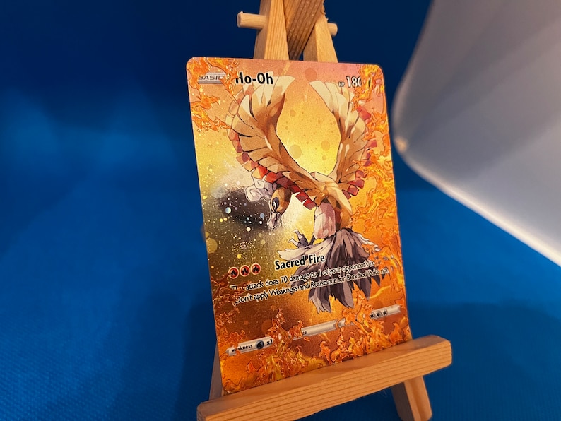 Pu&ograve; includere: Una carta da gioco di Pokemon dorata con Ho-Oh, un Pokemon uccello leggendario con un design arancione e giallo fiammeggiante. Il testo della carta recita "Sacred Fire: Questo attacco infligge 70 danni a 1 dei Pokemon del tuo avversario in gioco. Non applicare Debolezza e Resistenza ai Pokemon in panchina."