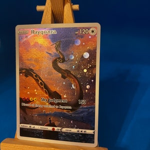 May include: A holographic Pokemon trading card featuring Rayquaza, a green dragon-like creature with a gold chain around its neck. The card has a background of a starry sky with a sunset in the distance. The card text reads "Sky Judgment: Discard all Energy attached to Rayquaza 150".
