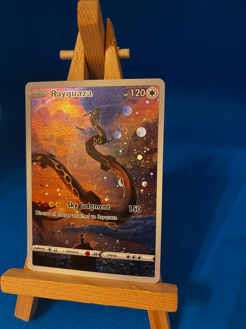May include: A holographic Pokemon trading card featuring Rayquaza, a green dragon-like creature with a gold chain around its neck. The card has a background of a starry sky with a sunset in the distance. The card text reads "Sky Judgment: Discard all Energy attached to Rayquaza 150".