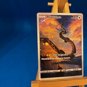 May include: A Rayquaza Pokemon card with a holographic finish. The card features a gold and black illustration of Rayquaza against a background of a sunset sky. The card has a HP of 120 and an attack called Sky Judgment that does 150 damage. The card text reads "Discard all Energy attached to Rayquaza."