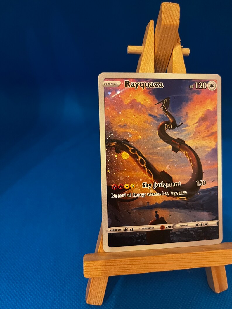 May include: A Rayquaza Pokemon card with a holographic finish. The card features a gold and black illustration of Rayquaza against a background of a sunset sky. The card has a HP of 120 and an attack called Sky Judgment that does 150 damage. The card text reads "Discard all Energy attached to Rayquaza."