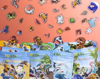 May include: A collection of colorful dinosaur stickers scattered above four Paleorium booster packs labeled Desert, Forest, Ocean, and Swamp. The stickers feature various prehistoric creatures, fossils, and plants in a variety of poses and colors.