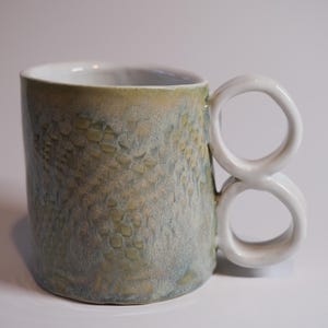 May include: A ceramic mug with a green and blue glaze and a unique double-loop handle.