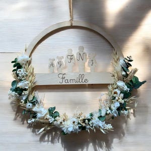 Peut inclure: Une couronne en bois avec une découpe d'une famille avec le mot "Famille" écrit en dessous. La couronne est décorée de fleurs séchées et de verdure.