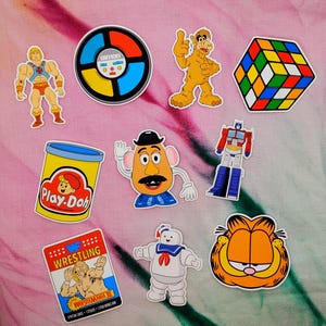 May include: A collection of colorful stickers featuring pop culture characters and icons. Includes He-Man, a Simon game, Alf, a Rubik's Cube, Play-Doh, Mr. Potato Head, Optimus Prime, a wrestling card, the Stay Puft Marshmallow Man, and Garfield.