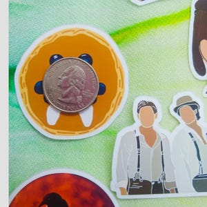 May include: A quarter coin rests on a round orange sticker with blue accents and white teeth-like shapes. Other stickers include illustrations of people and a raven silhouette against a red moon.