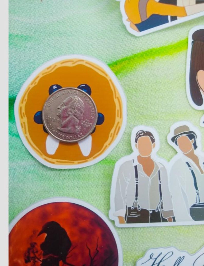 May include: A quarter coin rests on a round orange sticker with blue accents and white teeth-like shapes. Other stickers include illustrations of people and a raven silhouette against a red moon.