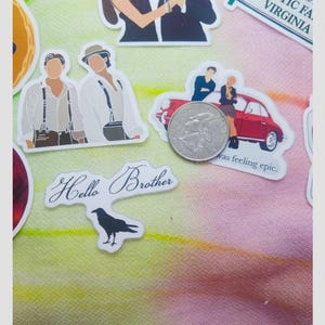 May include: A collection of stickers featuring illustrations of people, a red car, and the text "Hello Brother." The stickers are on a colorful, patterned background. A quarter is included for scale.