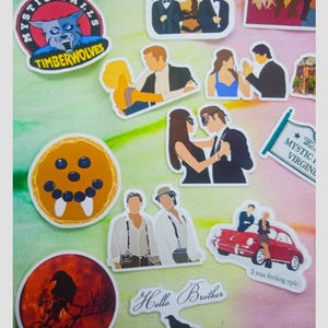 May include: A collection of stickers featuring various designs. Included are stickers with the text "Mystic Falls TimberWolves", a raven silhouette, and the phrase "Hello Brother". Other stickers depict cartoon figures and scenes.