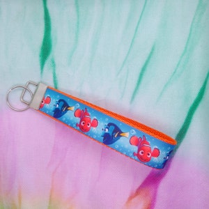 May include: A Finding Nemo themed keychain featuring Nemo and Dory characters on a blue background with bubbles. The keychain has an orange border and a silver key ring. The background is a mix of pink, green, and white.