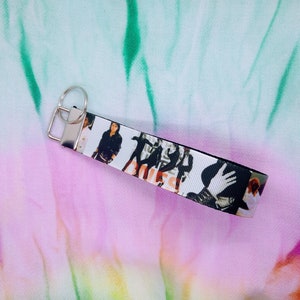 May include: A white and black wristlet keychain featuring a repeating pattern of a group of people. The keychain has a silver-colored metal clasp and key ring. The background is a tie-dye pattern of pink, green, and white.