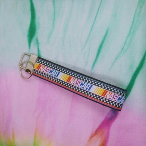 Puede incluir: Un llavero NASCAR en blanco y negro con la palabra "NASCAR" en blanco, azul y rojo. El llavero tiene un anillo y un cierre de metal plateado. El fondo es un patrón tie-dye de color rosa, verde y blanco.