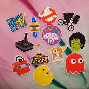 May include: A collection of colorful stickers featuring pop culture icons. Includes a Ghostbusters logo, ET on a bike, a Pac-Man character, a Gremlin, and a joystick. Other stickers include the MTV logo and a "Book It!" design.