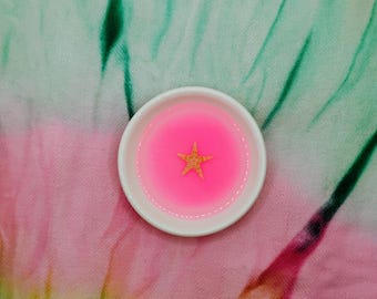 May include: A small, white, round dish filled with bright pink liquid and a gold star. The dish is centered on a colorful, tie-dye background of pink, green, and white.