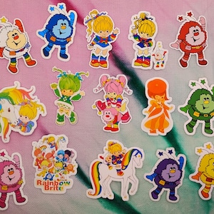 May include: A collection of colorful Rainbow Brite stickers featuring various characters and scenes. The stickers showcase characters with vibrant hair, outfits, and whimsical designs, including a rainbow-colored unicorn and the text "Rainbow Brite".