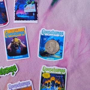 K&ouml;nnte beinhalten: Eine Sammlung von Goosebumps-Sammelkarten-Aufklebern mit verschiedenen Buchcovern. Die Aufkleber zeigen lebendige Illustrationen und Titel wie "Horrorland", "The Haunted Mask" und "Attack of the Jack-O-Lanterns". Eine M&uuml;nze ist enthalten.