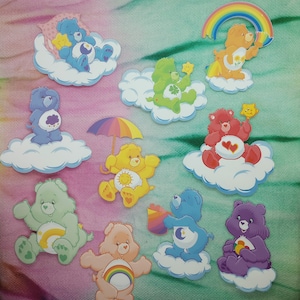 May include: A collection of colorful Care Bear stickers. The stickers feature various bears in different poses, each on a cloud. The bears are in shades of blue, green, yellow, red, and purple, with rainbow and star accents.