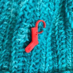 Peut inclure: Un marqueur de point en plastique rouge en forme de chaussette, placé sur un tissu tricoté turquoise.