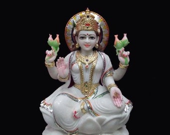 May include: White marble statue of a Hindu goddess sitting with her legs crossed. She is wearing a white dress and a gold crown. She is holding a flower in each hand. The statue is decorated with gold and red paint.