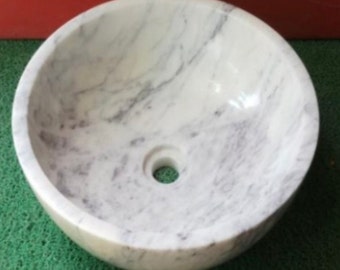 May include: A round vessel sink crafted from white marble with natural grey veining throughout. The basin features a smooth, polished interior surface and a centered circular drain hole at the base, showcasing the unique stone pattern and texture.