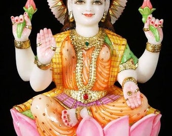 May include: A white marble statue of a Hindu goddess, Lakshmi, sitting on a pink lotus flower. She is adorned with gold jewelry and a yellow and green sari. She holds a lotus flower in each hand.