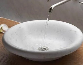 May include: A round white marble vessel sink with subtle grey veining, featuring a polished finish and a central metal drain. A silver faucet is positioned above the basin, with a steady stream of water flowing directly into the center drain.