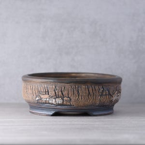 May include: A round ceramic bonsai pot with a textured, cracked brown exterior finish that resembles tree bark. The rim and the base of the pot are smooth and dark, providing a contrast to the rough, weathered appearance of the main body.