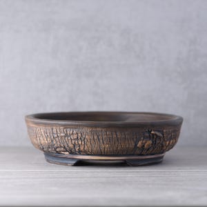 May include: A shallow, round ceramic bonsai pot with a dark brown, textured exterior that mimics the appearance of tree bark. The rim is smooth and dark, while the base features a raised footed design, providing a stable foundation for the vessel.