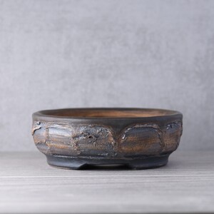 以下が含まれることがあります： 暗く風化した仕上げの丸く背の低い陶器製の盆栽鉢。外側は深い茶色とチャコールグレーの色調で、質感のあるスカラップ模様が施されており、内側は滑らかな自然な粘土色の表面が見えています。