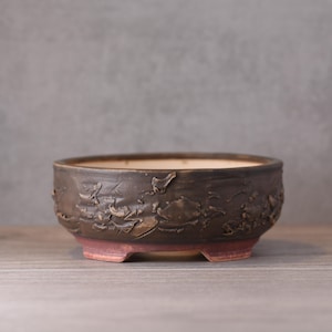 Könnte beinhalten: Ein dunkelbrauner Bonsai-Topf aus Keramik mit strukturierter Außenseite und rotbraunem Sockel. Der Topf hat eine breite, flache Form und steht auf drei kleinen Füßen. Der Innenraum ist hellbeige.