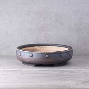 Könnte beinhalten: Eine flache, runde Bonsaischale aus Keramik mit einer dunklen, verwitterten Oberfläche in Anthrazit- und Kupfertönen. Der äußere Rand ist mit einer Reihe gleichmäßig verteilter, erhabener runder Noppen verziert. Das Innere ist hell und unglasiert.
