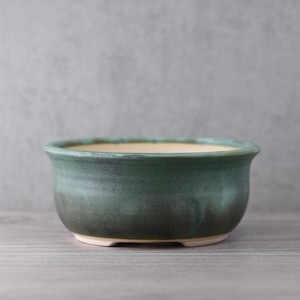 May include: A round, ceramic bonsai pot with a dark teal glaze. The pot has a wide, shallow shape with a slightly flared rim and a cream-colored interior. The exterior glaze transitions from dark teal to a deeper, almost black color at the base.