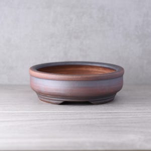 Op de afbeelding: Een ronde, lage bonsai-pot van keramiek met een matte, aardebruine afwerking. De buitenkant is voorzien van subtiele, donkergrijze horizontale banden rond het midden en een dikke, afgeronde rand. De basis heeft kleine, geïntegreerde voetjes.