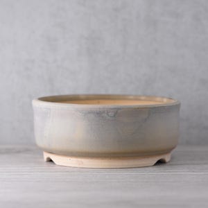 May include: A round, ceramic bonsai pot with a light blue and beige glaze. The pot has a low profile with a wide opening and sits on three small feet. The interior has a light beige color, and the exterior has a gradient of blue and beige.