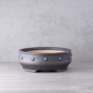 Könnte beinhalten: Ein runder, flacher Bonsai-Topf aus Keramik mit einer dunklen, matten anthrazitfarbenen Oberfläche und einer Reihe kleiner, erhabener kreisförmiger Noppen, die gleichmäßig um den äußeren Rand verteilt sind. Das Innere ist beige.