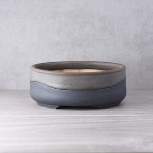 May include: A round, low-profile ceramic bonsai pot featuring a two-tone glaze. The upper portion is a muted, earthy grey, while the lower section is a deep, matte charcoal blue. The interior reveals a light, natural clay finish with a wide opening.