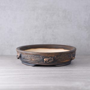 May include: A round, shallow ceramic bonsai pot with a dark, textured exterior that mimics the appearance of charred wood or rough bark. The interior of the pot features a smooth, light beige finish, and the base is supported by small feet.