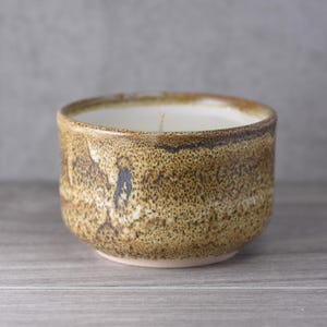 Könnte beinhalten: Eine handgefertigte Kerze aus Keramik in einem runden, strukturierten, braunen Gefäß. Die Kerze hat einen weißen Wachsinnenraum und einen einzelnen Docht. Die Außenseite des Gefäßes hat ein gesprenkeltes Aussehen mit dunkleren braunen und blauen Akzenten.