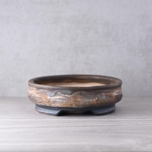 May include: A round, shallow ceramic bonsai pot with a textured, earthy brown and charcoal grey exterior. The rim is smooth and dark, while the base features small, integrated feet. The interior of the pot is a lighter, neutral beige tone.