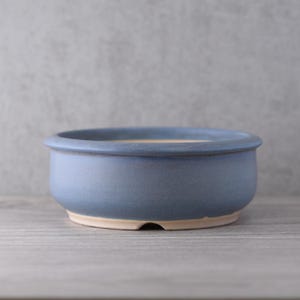 Könnte beinhalten: Ein runder, blauer Bonsai-Topf aus Keramik mit hellbeigem Innenraum. Der Topf hat ein breites, flaches Design mit einem leicht erhöhten Rand. Die Außenseite hat eine matte Oberfläche und die Basis ist unglasiert. Der Topf ist für Bonsai-Bäume konzipiert.