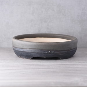 May include: A shallow, oval-shaped ceramic bonsai pot featuring a two-tone finish. The upper rim is a smooth, matte charcoal gray, while the lower exterior displays a textured, dark navy blue glaze with visible brushstroke details and small feet.