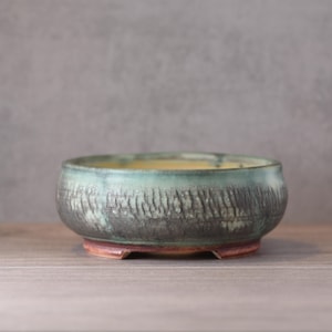 Pode incluir: Uma tigela de cerâmica artesanal com um exterior cinza escuro texturizado e uma borda verde clara. A tigela tem uma base marrom avermelhada com três pequenos pés. O interior tem um tom amarelo. Esta tigela decorativa é adequada para plantas ou como peça decorativa.