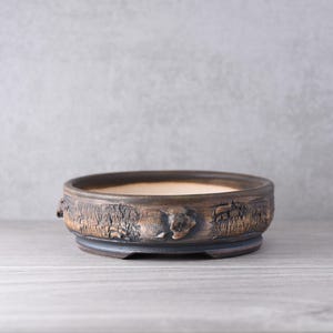 Peut inclure: Un pot à bonsaï rond et peu profond en céramique, doté d'un extérieur brun foncé texturé imitant l'écorce d'un arbre. L'intérieur du pot est d'une couleur beige lisse et la base présente un rebord circulaire foncé légèrement surélevé.